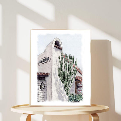 Framed watercolor art of Old Town Saloon San Diego with cactus and adobe-style architecture, displayed on a round wooden table. Southwestern wall decor inspired by historic Old Town San Diego charm