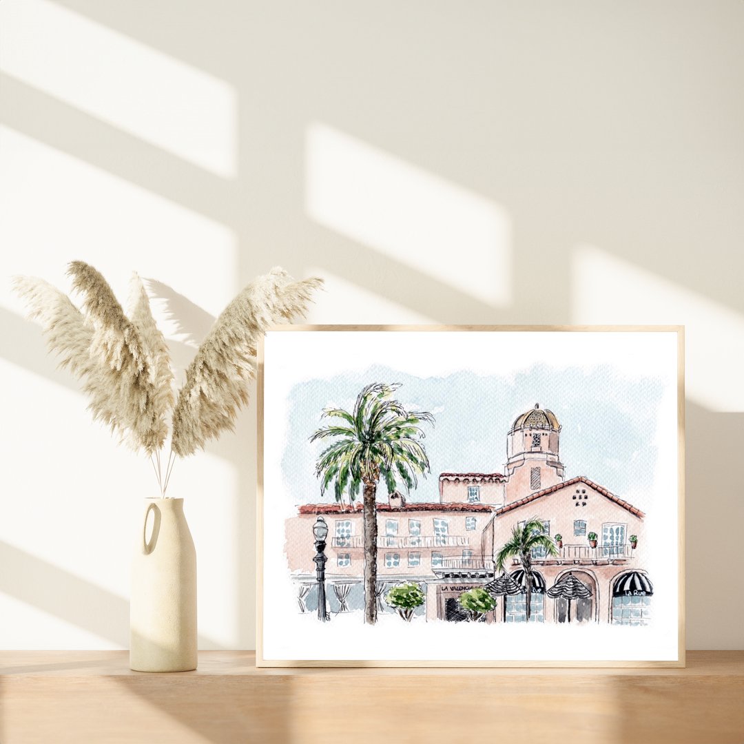 Framed watercolor painting of La Valencia Hotel, a historic pink building in La Jolla, California, displayed on a wooden shelf with dried pampas grass in a ceramic vase. The artwork highlights the hotel’s Mediterranean architecture, dome tower, and surrounding palm trees, with natural sunlight casting soft shadows