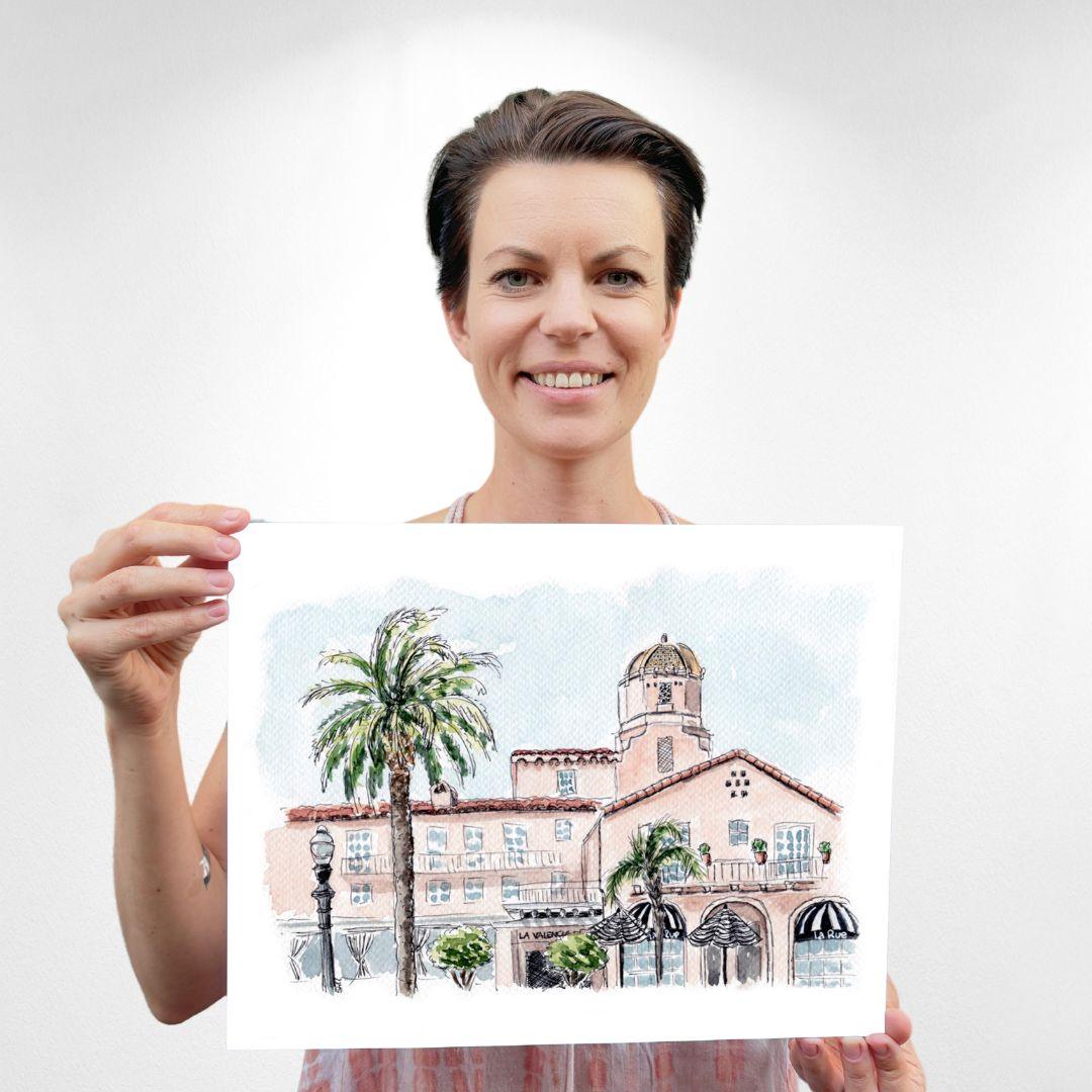 Artist Sara from Swil Arts holding her original watercolor print of La Valencia Hotel, known as 'The Pink Lady' in La Jolla, featuring vibrant palm trees and Spanish architecture. Perfect wall art for California lovers. Handcrafted watercolor art for home decor. Swil Arts watercolor print