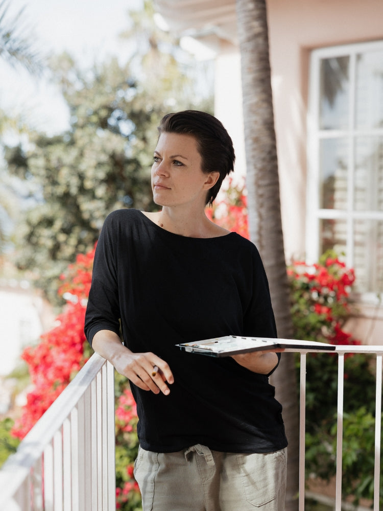 Sara, the artist behind Swil Arts, looking to the horizon and holding a watercolor palette. She is wearing a black shirt. The background is blurred but you can notice a bougainvillea. 