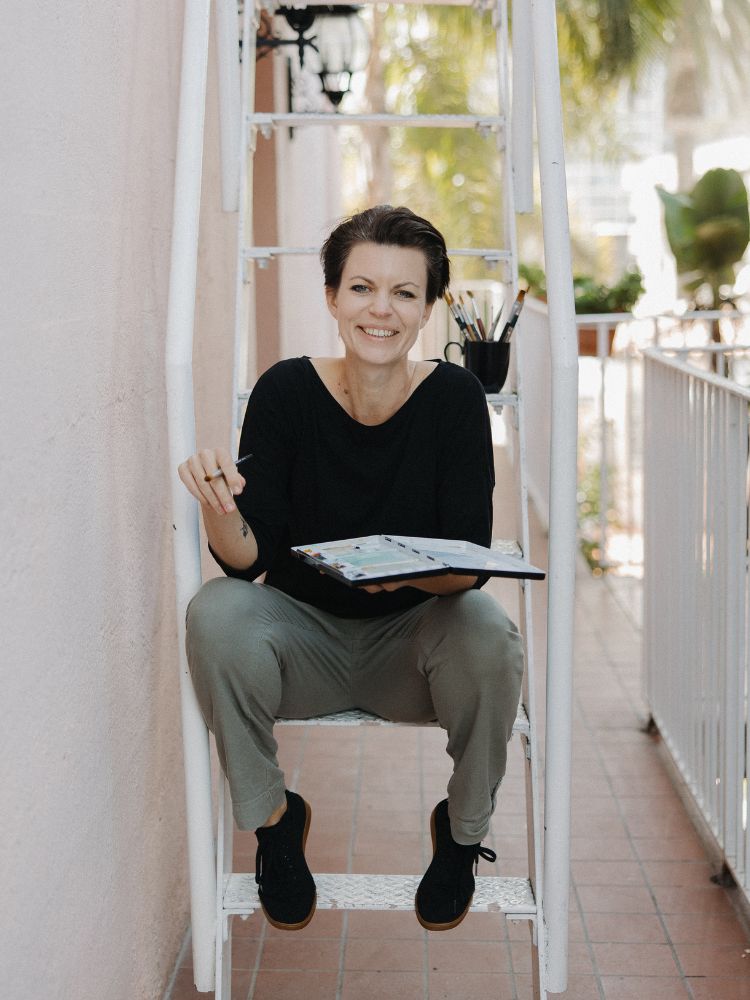 Sara, the artist behind Swil Arts, sitting on a ladder with a big smile on her face. She is wearing a black shirt and green pants and is holding a watercolor palette