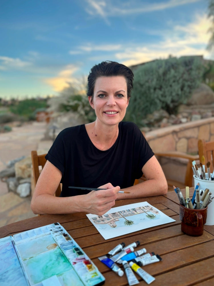 Sara, the artist behind Swil Arts is looking at the camera with a big smile on her face. She is holding a paintbrush and is giving the final touches to one of her art pieces. You can see paintbrushes, watercolor tubes and palette. The background is blurred but you can see a beautiful blue sky