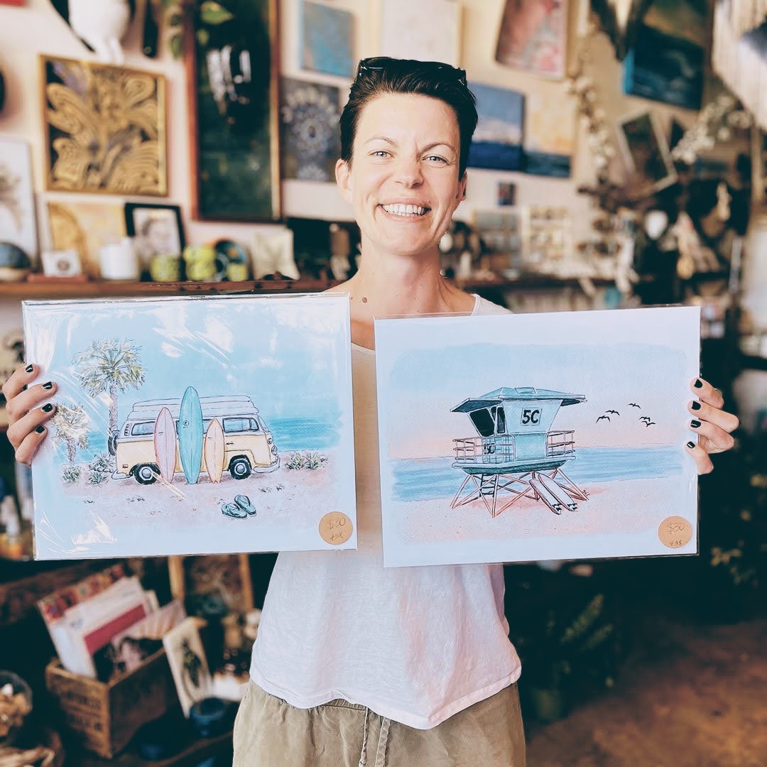 Sara, the artist behind Swil Arts is holding two of her prints. One is a surf van with palm treats and the other is a lifeguard tower in a beautiful sunset